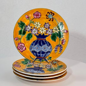 May include: A set of four yellow ceramic plates with a floral design. The plates feature a blue and white vase with a floral arrangement of pink, purple, and yellow flowers.