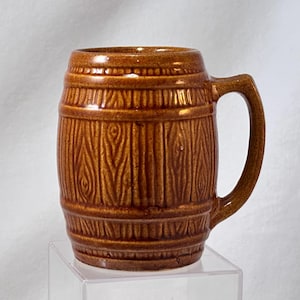 May include: A brown ceramic mug with a barrel design. The mug has a handle and is decorated with vertical lines and wood grain texture. The mug is approximately 5 inches tall.