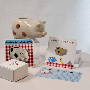 May include: A collection of pig-themed items, including a ceramic pig planter with brown spots, a pin, a card with a pig graphic, and a small box with "thank you" text. The items are arranged on a white surface.