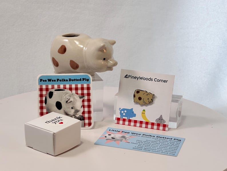 May include: A collection of pig-themed items, including a ceramic pig planter with brown spots, a pin, a card with a pig graphic, and a small box with "thank you" text. The items are arranged on a white surface.