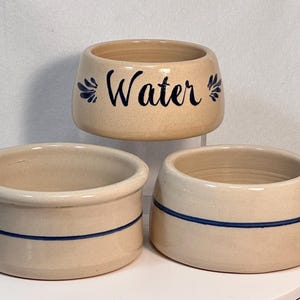 May include: Three beige ceramic bowls. The top bowl has the word "Water" in black script with blue floral accents. The other two bowls have a blue stripe around the middle. These bowls are suitable for pet food or water.