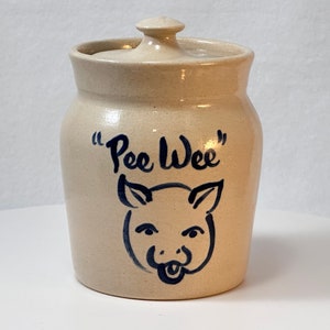 May include: A beige ceramic jar with a lid, featuring a blue and white illustration of a pig with the text "Pee Wee" on the front.