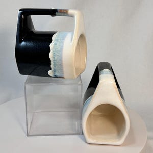 May include: Two ceramic mugs with a black and white glaze. The mugs have a unique shape with a handle that is part of the mug's body. The mugs are sitting on a clear acrylic surface.