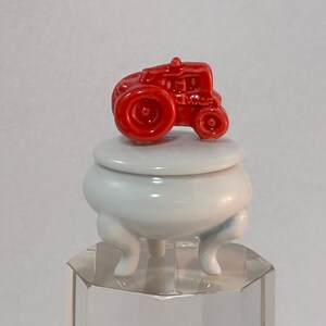 May include: A small red ceramic tractor figurine sits on top of a white ceramic lidded jar with three legs. The jar is on a clear glass base.