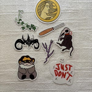 May include: A collection of stickers featuring various designs. Included are a gold-framed profile, a carving tool, a black and white abstract design, a plague doctor, a cartoon creature, a sprig of ivy, a purple lightning bolt, and a sticker with the text "JUST DON'T".