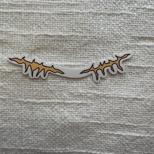 May include: A sticker featuring a stylized, curved design with a white background. The design is composed of a series of jagged, orange-yellow shapes, resembling a stylized eyebrow or a decorative element.