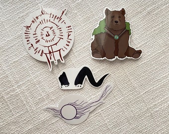 May include: Four stickers featuring a brown bear with a green cloak and a turquoise necklace, a black and white swirl, a black paw print, and a white circle with a black swirl and a white circle in the center.