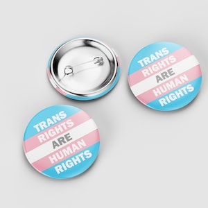 May include: Three circular badges with a light blue, white, and pink striped design. The text "TRANS RIGHTS ARE HUMAN RIGHTS" is printed on the front. One badge is face up, and another is face down, showing the pin back.
