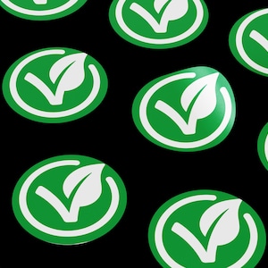 May include: A pattern of green and white circular stickers with a leaf and checkmark design. The stickers are on a black background.