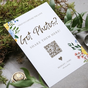 Puede incluir: Tarjeta blanca con borde floral y letras doradas. El texto dice "Help us capture the love Got Photos? Share them here!" con un código QR y el texto "Bridget Tori 06.03.2023".