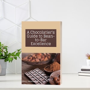 May include: A book titled "A Chocolatier's Guide to Bean-to-Bar Excellence" with a cover image featuring cocoa beans, chocolate, and cocoa powder. The book is on a white surface with a plant and decorative wall in the background.
