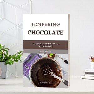 May include: Book cover with the title "Tempering Chocolate: The Ultimate Handbook for Chocolatiers" with a photo of melted chocolate in a bowl and a whisk. The book is published by Chocolate Den.