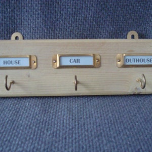 May include: A wooden key holder with three hooks and three brass labels. The labels read "HOUSE", "CAR", and "OUTHOUSE".