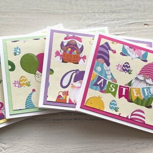 May include: Set of four Easter greeting cards featuring gnomes with Easter eggs, bunnies, and balloons. The cards have a light yellow background with colorful accents. The text on the cards reads "Hop" and "Easter".