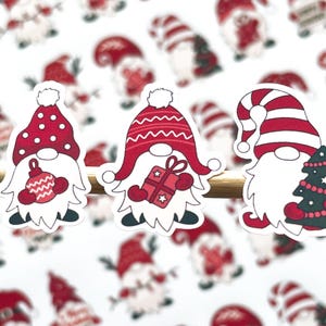 May include: Three red and white Christmas gnomes with different designs. One gnome is holding a red ornament, another is holding a gift box, and the third is holding a small Christmas tree.