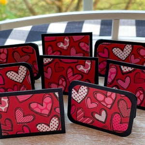 May include: Nine red and pink heart-shaped place cards with black borders. The hearts have different patterns, including polka dots, stripes, and swirls.