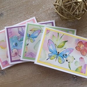 May include: Four blank greeting cards with watercolor butterfly designs. The cards are in shades of pink, purple, green, and yellow.