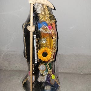 Peut inclure: Statue décorative d'une figure vêtue d'une robe avec un visage de crâne, tenant une faux. La figure est noire avec des bordures dorées et ornée d'embellissements colorés, dont un tournesol et des pièces. La statue mesure environ 30 cm de haut.