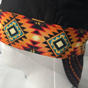 May include: A black welding cap featuring a wide band with a geometric pattern in orange, yellow, black, and turquoise. The fabric band is attached to the solid black crown with visible orange zigzag stitching along the top and bottom edges.