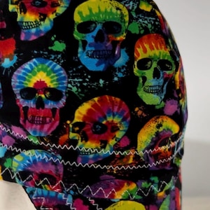 Può includere: Un berretto in tessuto con sfondo nero e un motivo ripetuto di teschi in vivaci colori tie-dye, tra cui giallo, verde, blu e rosa. Il berretto è realizzato con visibili cuciture a zigzag bianche lungo le giunture e i bordi.