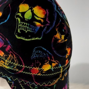 Può includere: Un berretto da saldatore nero con un motivo ripetuto di teschi in vivaci colori arcobaleno. Il tessuto è decorato con diverse file di cuciture a zigzag gialle a contrasto lungo il bordo inferiore, che aggiungono un dettaglio distintivo.