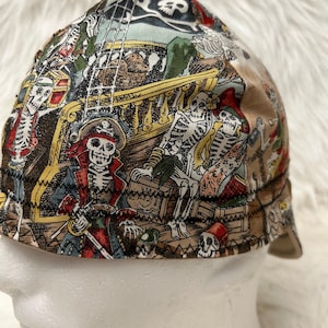 Puede incluir: Una gorra de tela multicolor con un estampado detallado de esqueletos vestidos de piratas en la cubierta de un barco. El diseño incluye una bandera pirata con una calavera, cajas de madera y aparejos de barco sobre un fondo beige y azul claro.