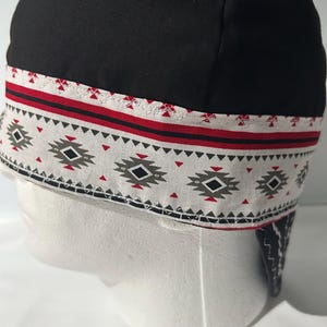 Może przedstawiać: Czarna czapka spawalnicza z szerokim pasem ozdobionym geometrycznym wzorem w kolorach czerwonym, czarnym i szarym na białym tle. Czapka posiada widoczne przeszycia wzdłuż krawędzi wzorzystego pasa oraz jednolitą czarną górną część.