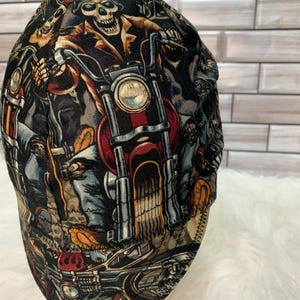 May include: A welding cap featuring a repeating graphic print of skeletons wearing goggles and riding motorcycles. The design includes black, red, and tan colors with detailed illustrations of bike engines, wheels, and handlebars across the fabric.
