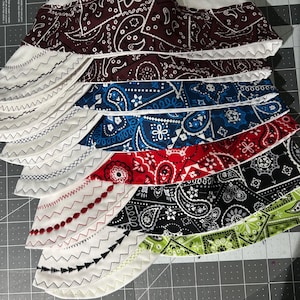May include: A stack of six fabric welding caps featuring a white crown and a patterned brim. The brims display various colors including maroon, blue, red, black, and lime green with paisley prints, accented by decorative zigzag stitching.