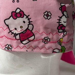 Może przedstawiać: Różowy czepek medyczny z powtarzającym się wzorem postaci Hello Kitty, żółtych pszczół i zielonych drzew. Tkanina jest ozdobiona dwoma rzędami jaskraworóżowych szwów zygzakowatych wzdłuż dolnej krawędzi, co stanowi dekoracyjne wykończenie.