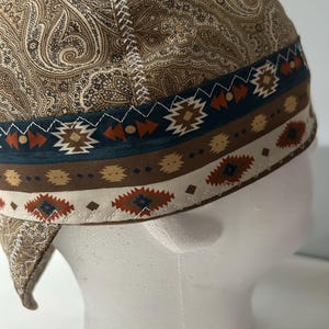 Puede incluir: Una gorra de soldador con un patrón de cachemira marrón en la corona y una banda decorativa con formas geométricas en tonos azul, naranja y crema. Los paneles de tela están unidos por costuras en zigzag blancas visibles en las uniones.