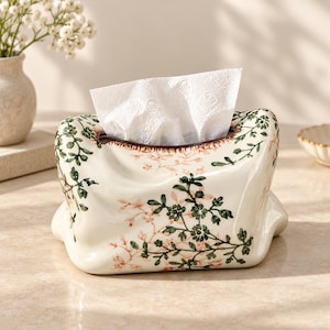 May include: A cream-coloured ceramic tissue box cover with a soft, organic, folded shape. The surface features a delicate botanical pattern of dark green leaves and light pink flowers. A white paper tissue is pulled through the top opening.