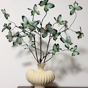Op de afbeelding: Een crèmekleurige keramische vaas bevat een decoratief arrangement van vlinder takken. De vlinders zijn in tinten groen en zwart, met delicate vleugelpatronen. De vaas heeft een unieke, afgeronde vorm met verticale ribbels.