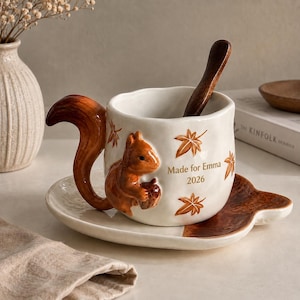 Puede incluir: Un juego de taza y plato de cerámica blanca con un asa tridimensional en forma de ardilla marrón y una figura de ardilla sosteniendo una bellota en el frente. La taza está decorada con motivos de hojas marrones y el texto Made for Emma 2026. Una cuchara de madera oscura descansa dentro.