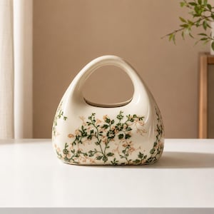 Puede incluir: Un jarrón de cerámica de color crema con una silueta redondeada similar a un bolso y un asa central recortada. La superficie exterior está decorada con un patrón delicado y repetitivo de pequeñas hojas verdes y flores color melocotón.