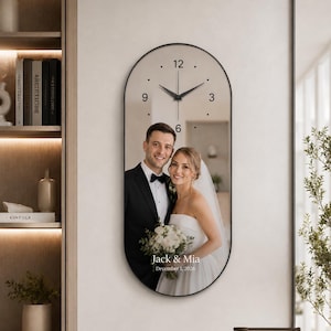 May include: An oval wall clock with a black frame featuring a printed photograph of a couple in wedding attire. The clock face has black hands, minimalist hour markers, and the numbers 3, 6, 9, and 12. Text reads Jack & Mia, December 1, 2026.