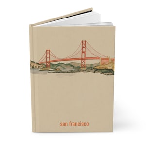Peut inclure: Un journal à couverture rigide avec une couverture beige. Il présente une illustration à l'aquarelle du Golden Gate Bridge à San Francisco. Le pont est rouge, et le texte "san francisco" est imprimé en orange en bas.