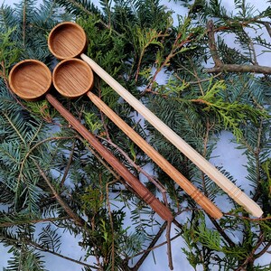 Può includere: Tre cucchiai di legno di diverse dimensioni e colori, con ciotole arrotondate e manici lunghi, sono disposti su un letto di rami di pino verdi.