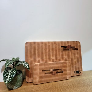 Peut inclure: Deux planches à découper en bois, l'une plus grande que l'autre, avec un motif en grille et un design décoratif. La plus petite planche est placée sur la plus grande.