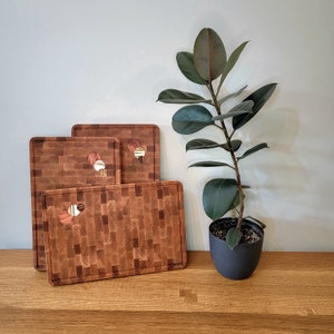 Peut inclure: Trois planches à découper en bois avec un motif en mosaïque unique. Les planches sont empilées les unes sur les autres et reposent sur une surface en bois. Une plante en pot se trouve en arrière-plan.