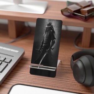 May include: A black and white phone stand with a samurai warrior image. The stand is made of metal and has a clear plastic holder for the phone.