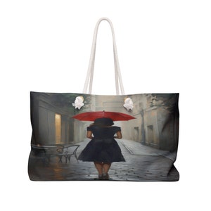 Puede incluir: Una bolsa de lona grande con una correa de lona blanca y una imagen impresa de una mujer caminando por una calle adoquinada con un paraguas rojo. La mujer lleva un vestido negro y la calle está bordeada de edificios.