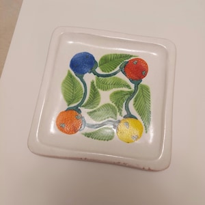 May include: A white ceramic square dish with a hand-painted design of four fruits and green leaves. The fruits are blue, red, orange, and yellow.