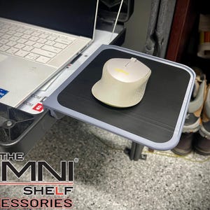 May include: A light-colored wireless computer mouse on a black mousepad. The mousepad is on a small, gray shelf attached to a laptop. The shelf is labeled "THE OMNI SHELF ACCESSORIES".