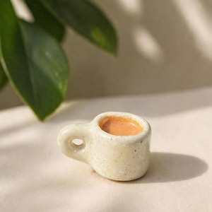 Könnte beinhalten: Eine kleine Kaffeetasse aus Keramik mit gesprenkelter, cremefarbener Glasur. Die Tasse enthält eine hellbraune Flüssigkeit, möglicherweise Kaffee oder Tee. Der Griff ist klein und abgerundet. Es ist ein dekorativer Artikel.