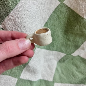 Puede incluir: Una taza pequeña de cerámica blanca con un líquido marrón en su interior. La taza tiene un esmalte moteado y una asa.