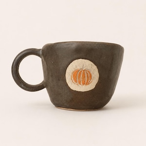 May include: A dark brown, hand-formed ceramic mug with an irregular, textured surface. Centered on the front is a circular, light-colored patch featuring a carved, orange-colored pumpkin illustration with visible vertical lines and a stem.