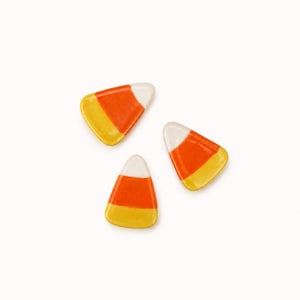 Könnte beinhalten: Drei Keramikornamente in Form von Candy Corn mit einer glatten, glänzenden Oberfläche. Jedes Stück zeigt ein horizontales dreifarbiges Muster mit einem weißen oberen Teil, einem leuchtend orangefarbenen Mittelband und einer gelben Basis.