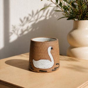 May include: A brown ceramic mug featuring a white swan design. The mug has a wide base and tapers upwards. The swan has a black beak and is set against black lines. A beige vase and foliage are in the background.