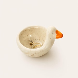 Puede incluir: Un joyero de cerámica con forma de ganso, con un acabado beige moteado y un pico naranja. El joyero contiene un anillo de oro con una gema grande y transparente. El joyero es un artículo decorativo.